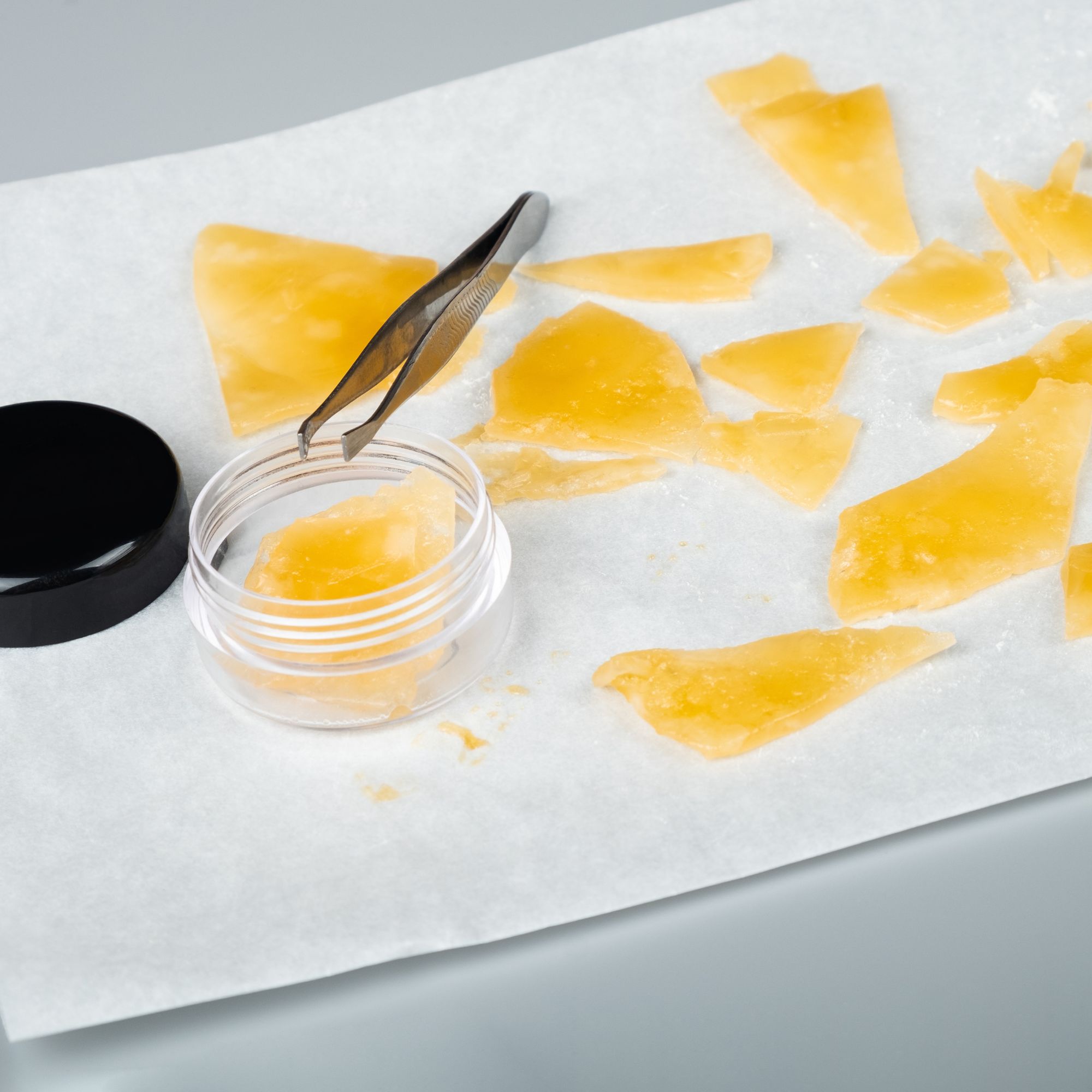 Pieces of cannabis shatter on wax paper, with tweezers picking up a piece.